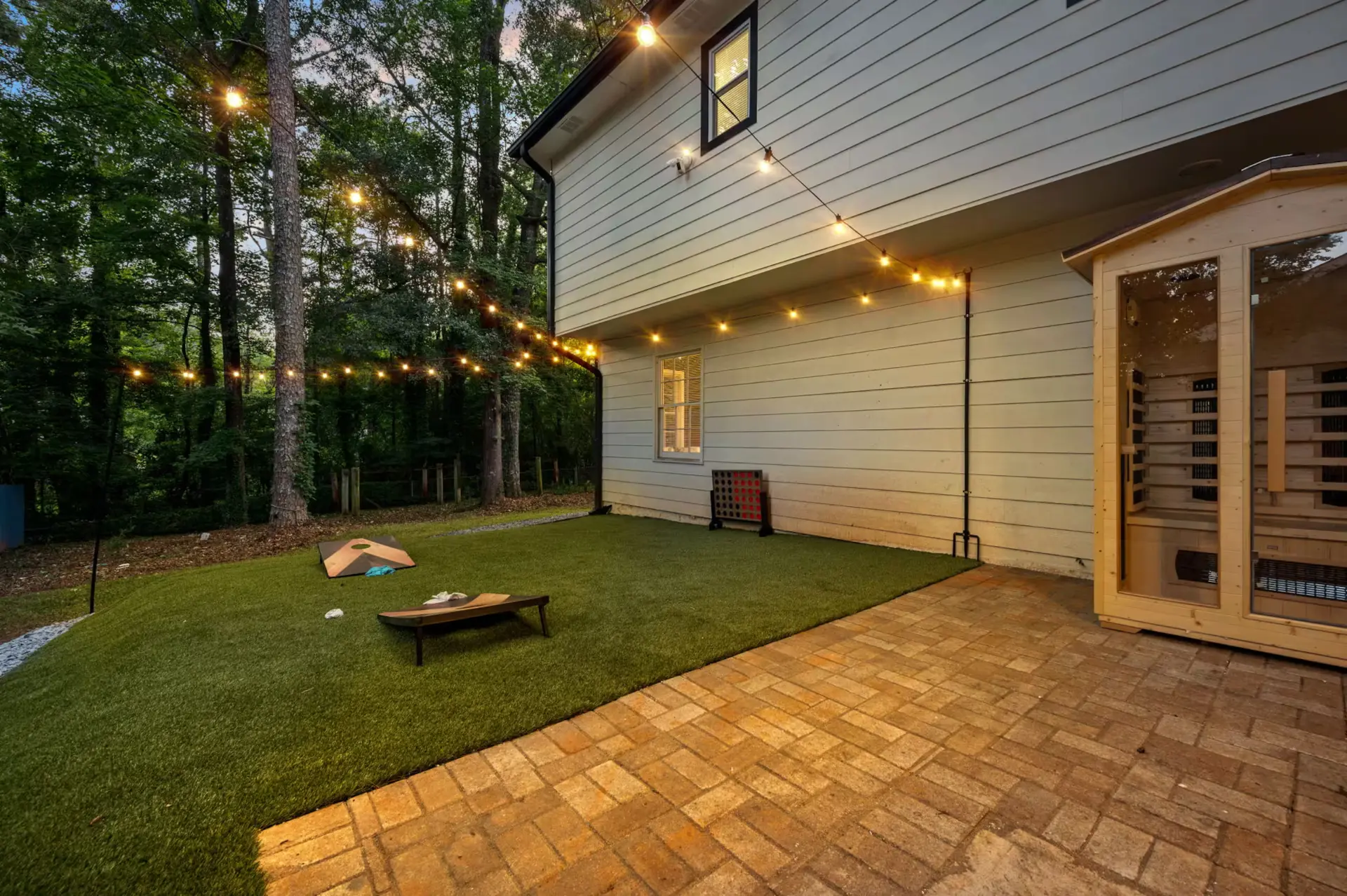 Backyard patio with cedar sauna, turf lawn, cornhole, and string lights at dusk