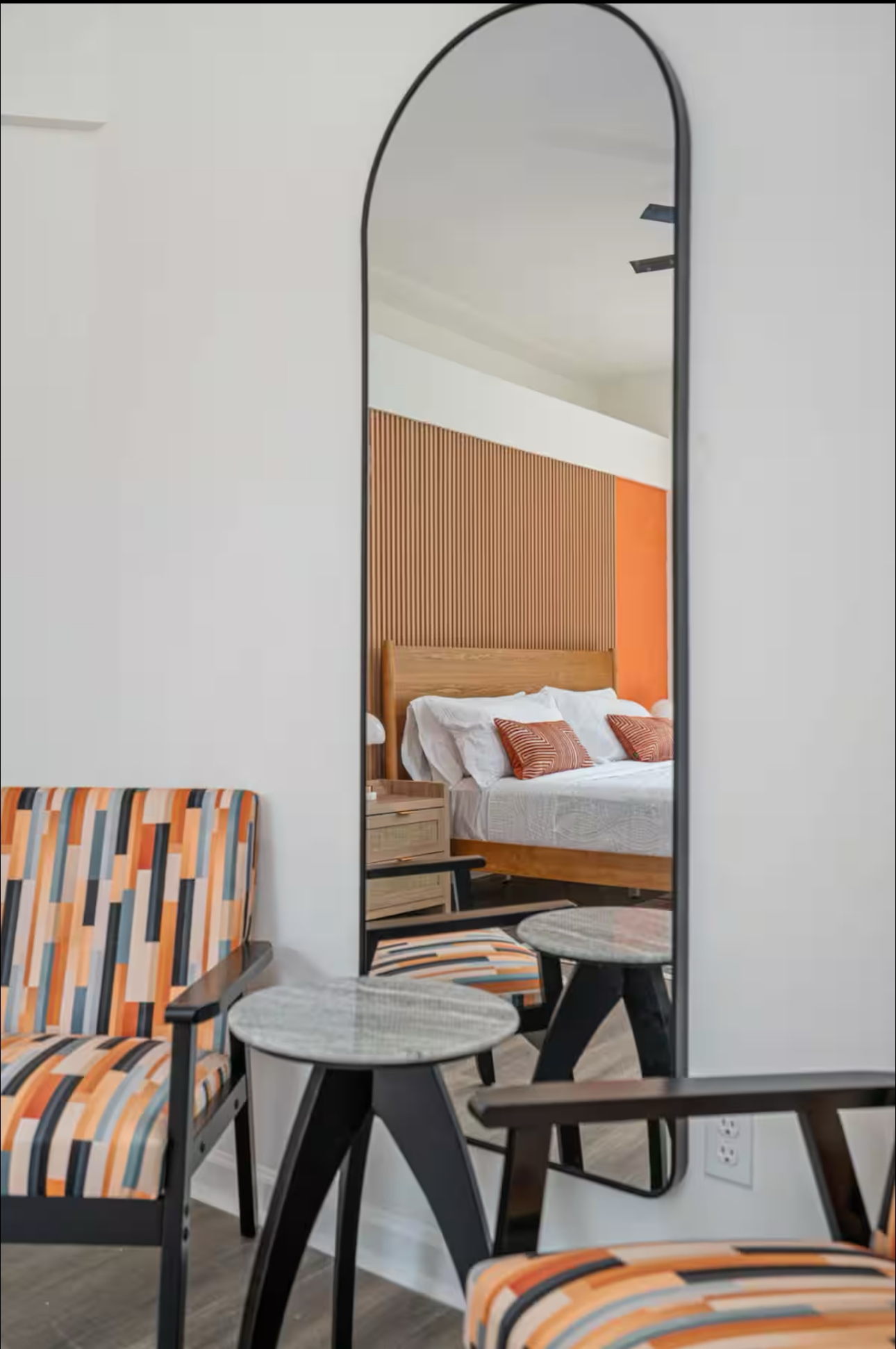 Arched full-length mirror reflecting the slat-wall bedroom beside striped accent chairs